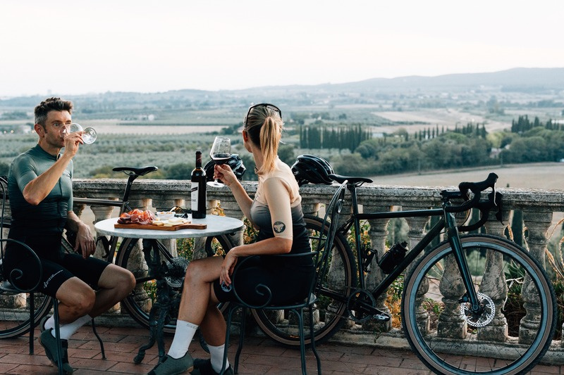 Two riders sharing wine on a terrace overlooking the Tuscan countryside